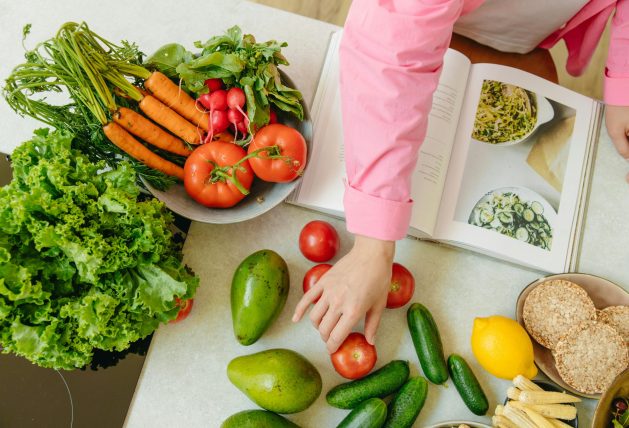 Top view of fresh vegetables and hands arranging them with a recipe book, promoting healthy eating.